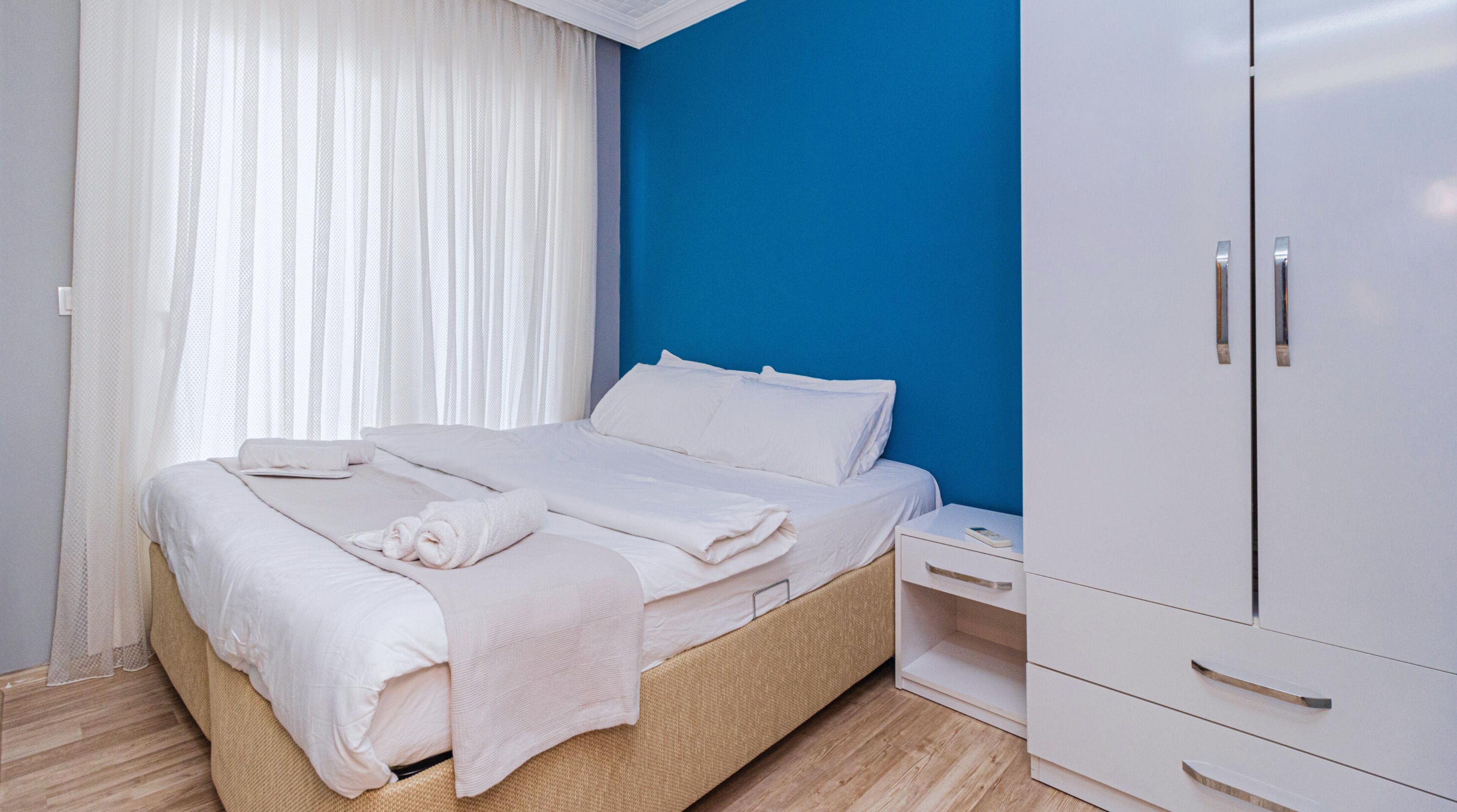 Een netjes opgemaakt bed met witte lakens en handdoeken, een nachtkastje met een boek, en een garderobe met zilveren handgrepen.