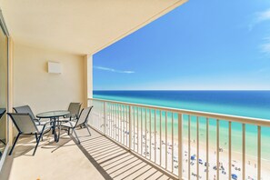Un balcon avec une table et des chaises, donnant sur une plage où l’on peut voir des gens et des parasols.