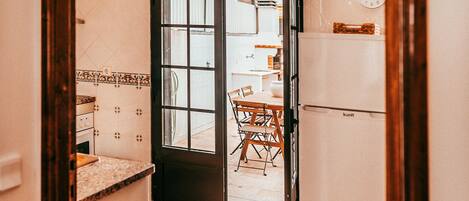 Une cuisine étroite avec un mur recouvert de carreaux, une porte en bois donnant sur un coin repas avec une table et des chaises, et un réfrigérateur situé à droite.