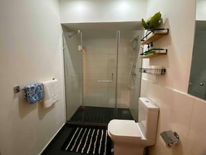 Un baño moderno con ducha envuelta en vidrio, inodoro y un pequeño estante con artículos de aseo.