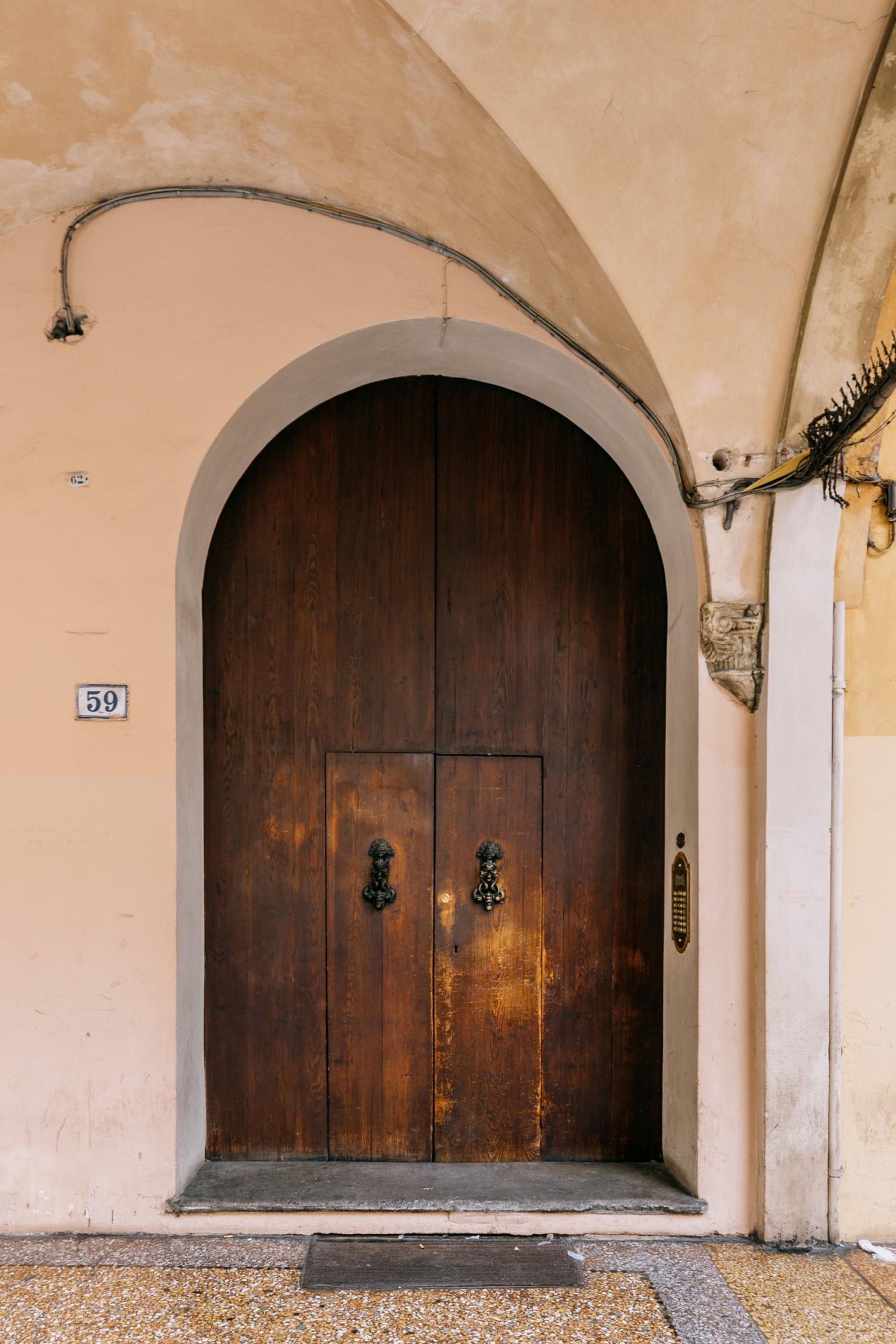 A large wooden double door with a metal handle, set in an arched doorway, and a number "59" sign on the wall to the left.