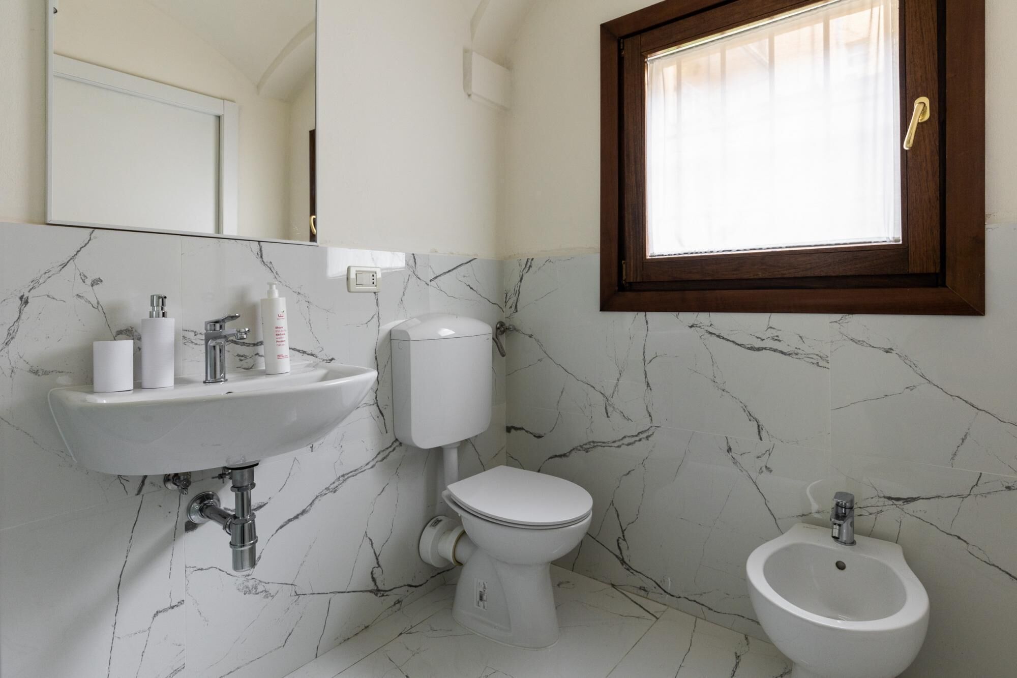 A bathroom with a marble sink, toilet, bidet, and a window with curtains.