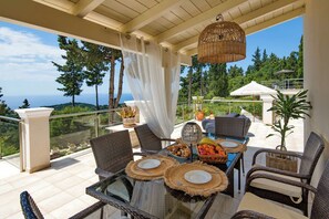 Un espacio comedor al aire libre iluminado por el sol, con mobiliario de mimbre, una mesa preparada para dos personas y vista al mar.
