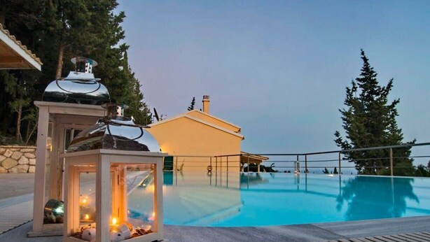 Une piscine avec une terrasse en bois, un petit bâtiment doté d’une cheminée et une lanterne allumée.