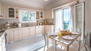 Une cuisine moderne avec des meubles blancs, un coin repas doté d’une table en verre et une vue sur l’extérieur grâce à de grandes fenêtres.