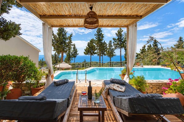 Zona junto a la piscina con dos tumbonas, una mesa con una copa de vino tinto y vistas a una piscina y árboles.