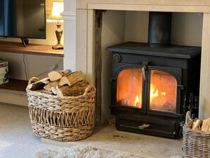 Une pièce chaleureuse avec un poêle à bois, un panier en osier rempli de bois de chauffage et une lampe posée sur une étagère.