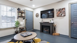A modern living room with a round table, a fireplace, a mounted TV, and a large window with blinds. - Take a trip to the Industrial Age in this centrally-located townhome. (Springdale)
