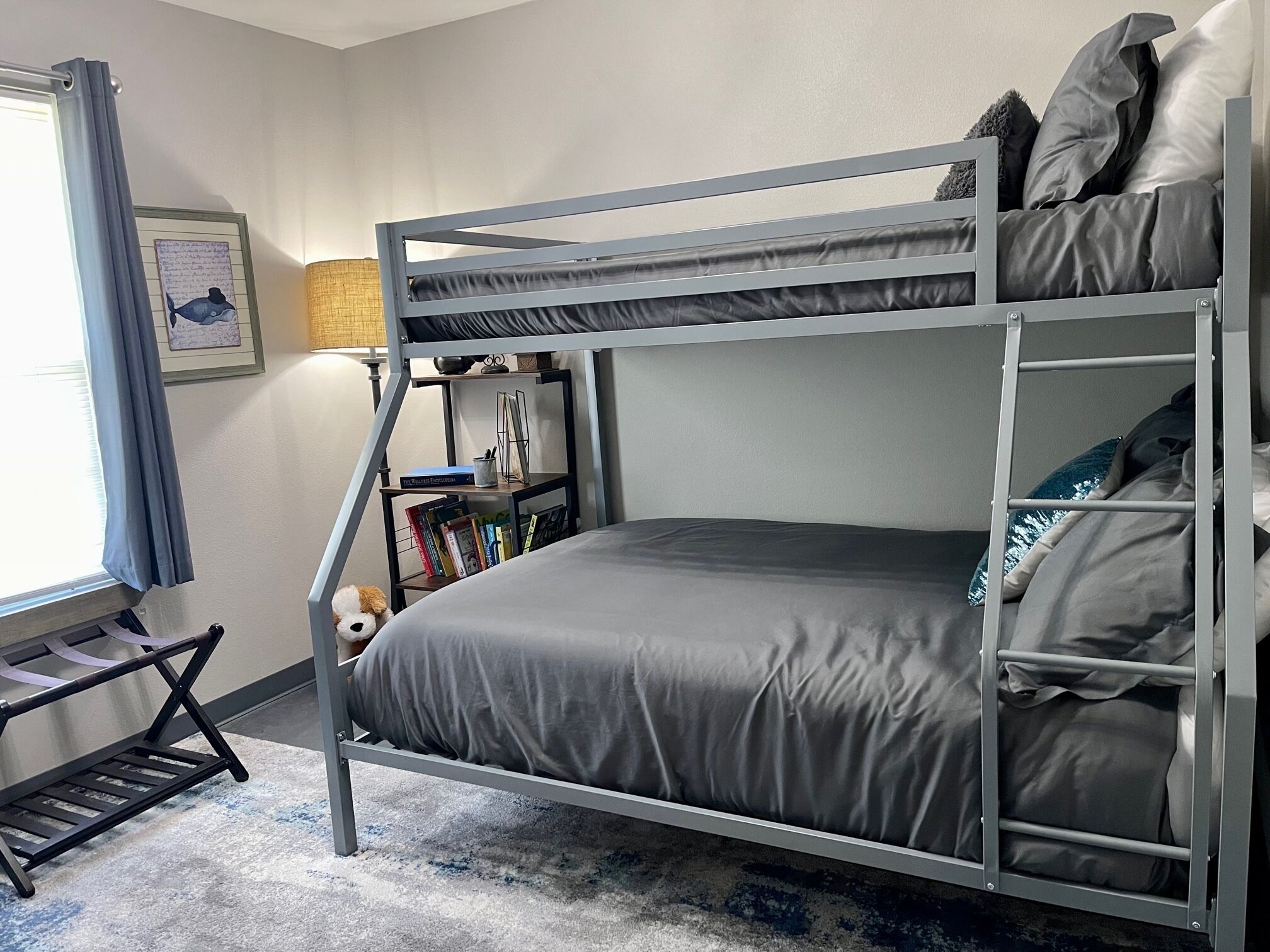 A bunk bed with a grey mattress, a small shelf with books, a lamp, and a window with curtains.
