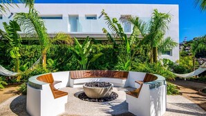 A modern outdoor seating area with a circular fire pit, surrounded by white benches with wooden accents, set against a backdrop of lush greenery and a white building. - Simply Splendid - Centrally Located, Amazing Pool/Gym! (Puerto Vallarta)
