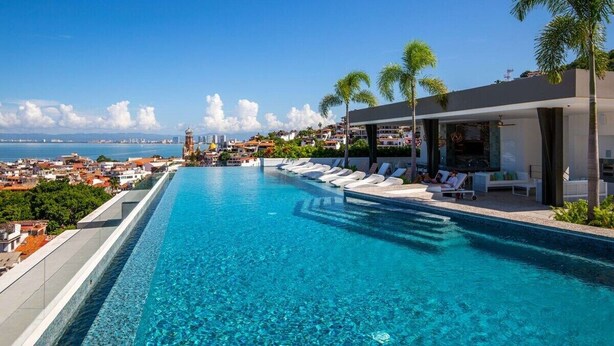 A clear blue pool with lounge chairs and palm trees, overlooking a coastal cityscape. - Simply Splendid - Centrally Located, Amazing Pool/Gym! (Puerto Vallarta)