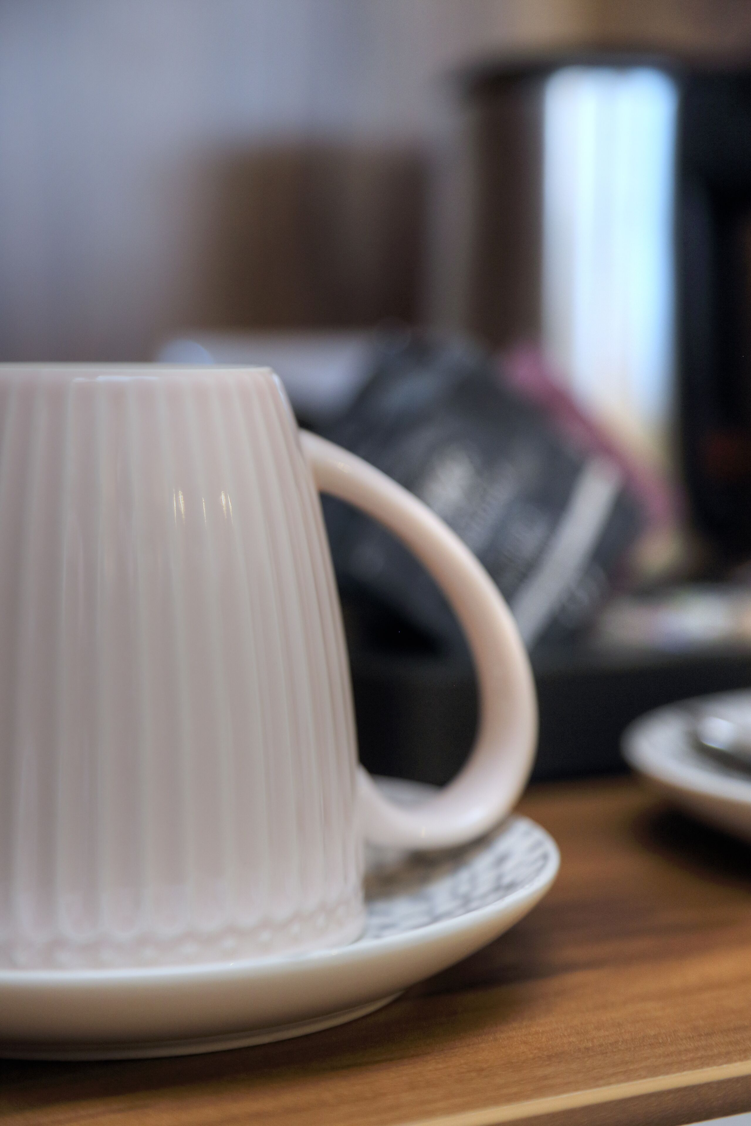 Une tasse en céramique blanche posée sur une soucoupe, une assiette à motifs noir et blanc, et un arrière-plan flou avec un objet métallique.