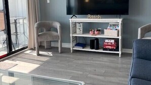 Una sala de estar con una televisión de pantalla plana, un mueble blanco para equipo multimedia, dos sillones y una mesa de centro de vidrio.