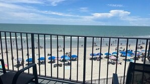 Vista a la playa desde un balcón con barandilla, sombrillas y gente en la arena.