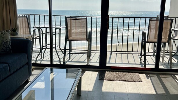 Un balcón con mesa de vidrio, sillas y sofá, con vista a una playa con olas y cielo despejado.