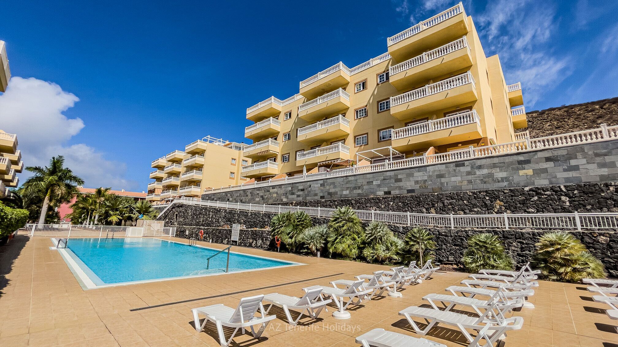 Um hotel com piscina, espreguiçadeiras brancas e um muro de pedra.