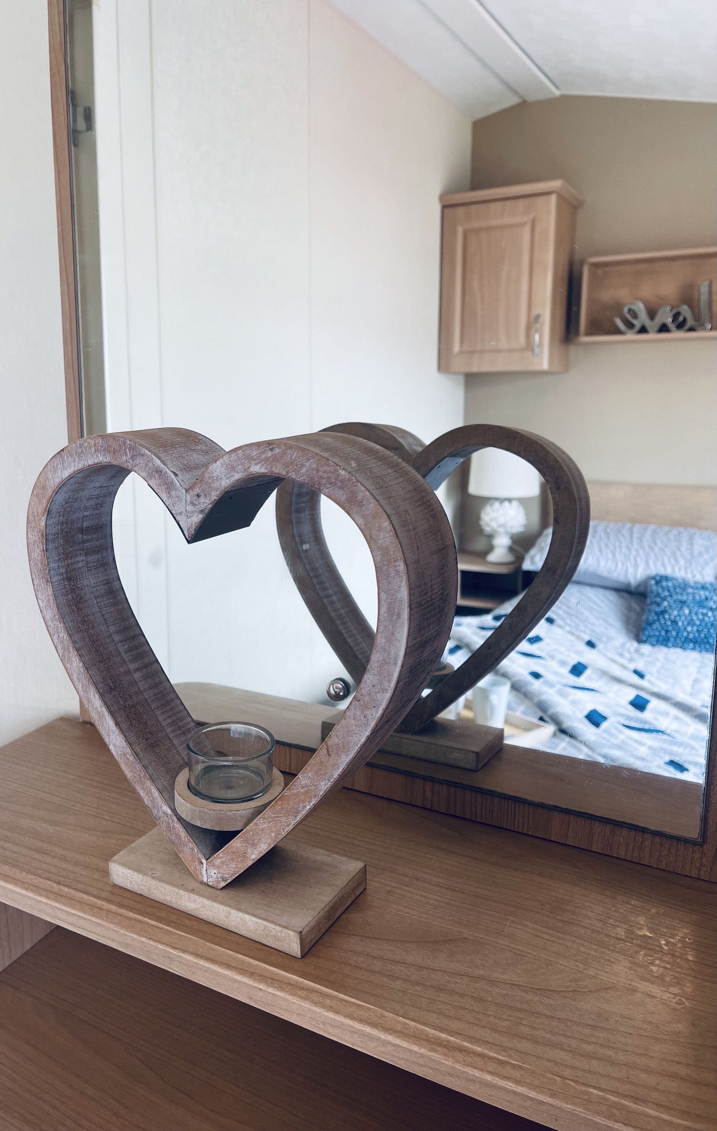 A wooden heart-shaped candle holder on a wooden table with a mirror reflecting a bedroom.