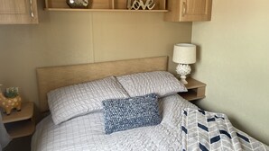 A neatly made bed with a quilted headboard, a bedside lamp, and a wooden shelf above holding decorative items. - Charming 3-bed Static Caravan in Porthcawl (Porthcawl)