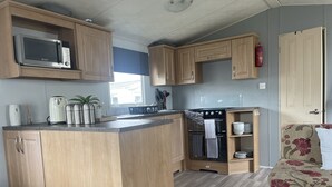 A compact kitchen with wooden cabinets, a microwave, a stove, and a sink. - Charming 3-bed Static Caravan in Porthcawl (Porthcawl)