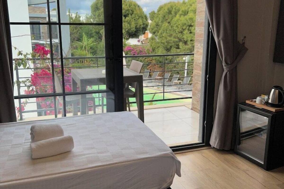 A bedroom with a bed, a small table with a kettle, and a view of a balcony with outdoor furniture.