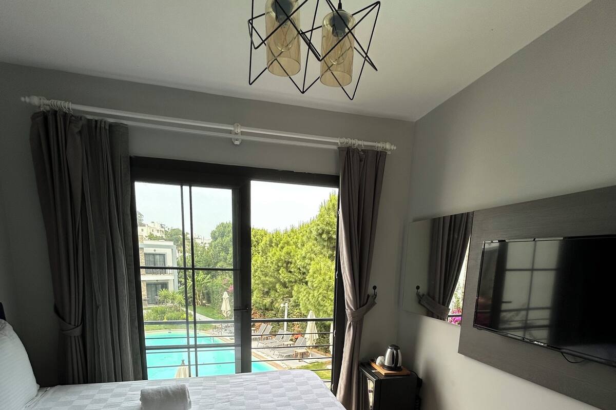 A hotel room with two beds, a TV, a window with a view of a pool and greenery, and a modern chandelier.