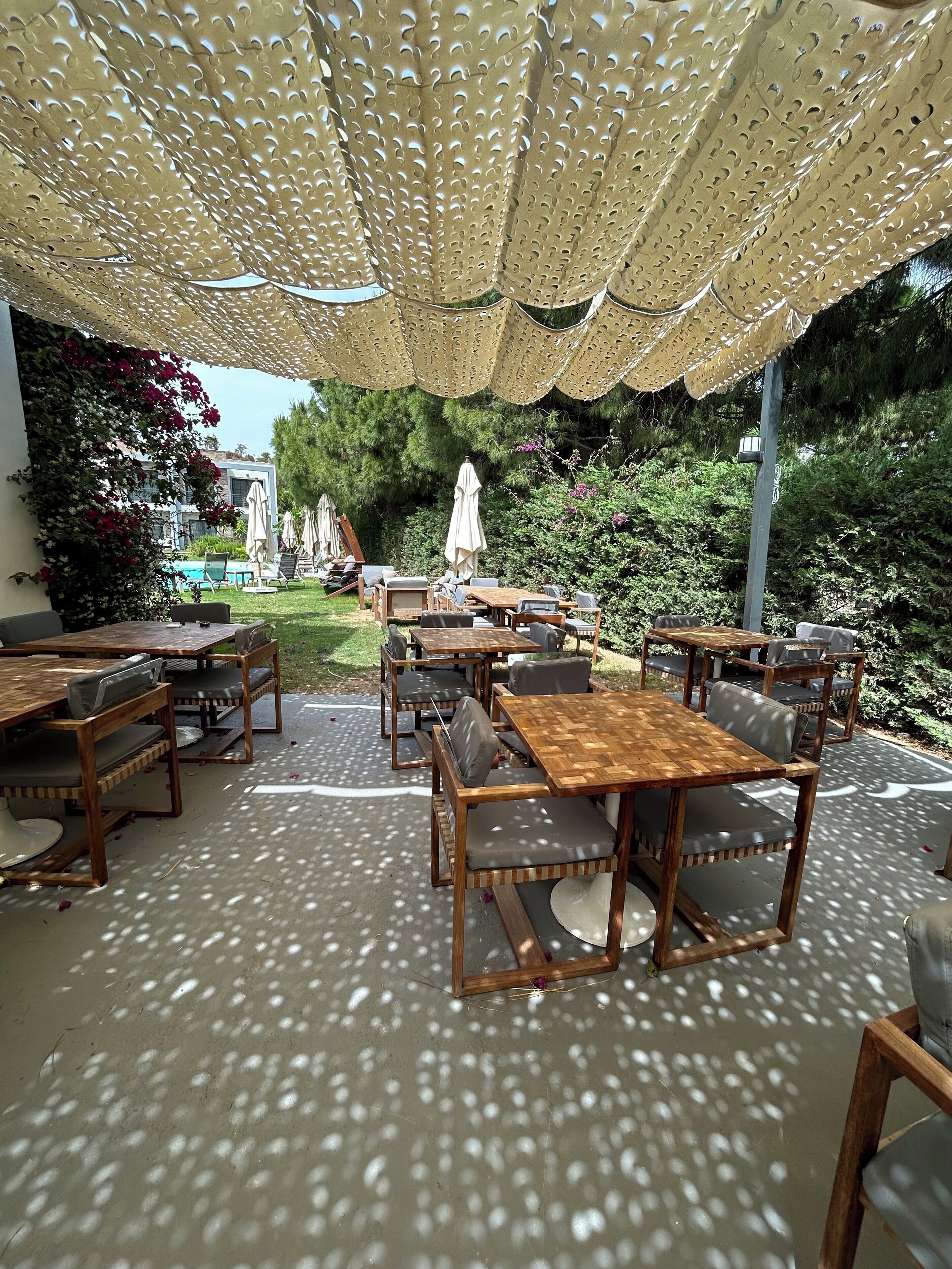 Un patio al aire libre con mesas y sillas de madera, una pérgola en lo alto y una piscina al fondo.