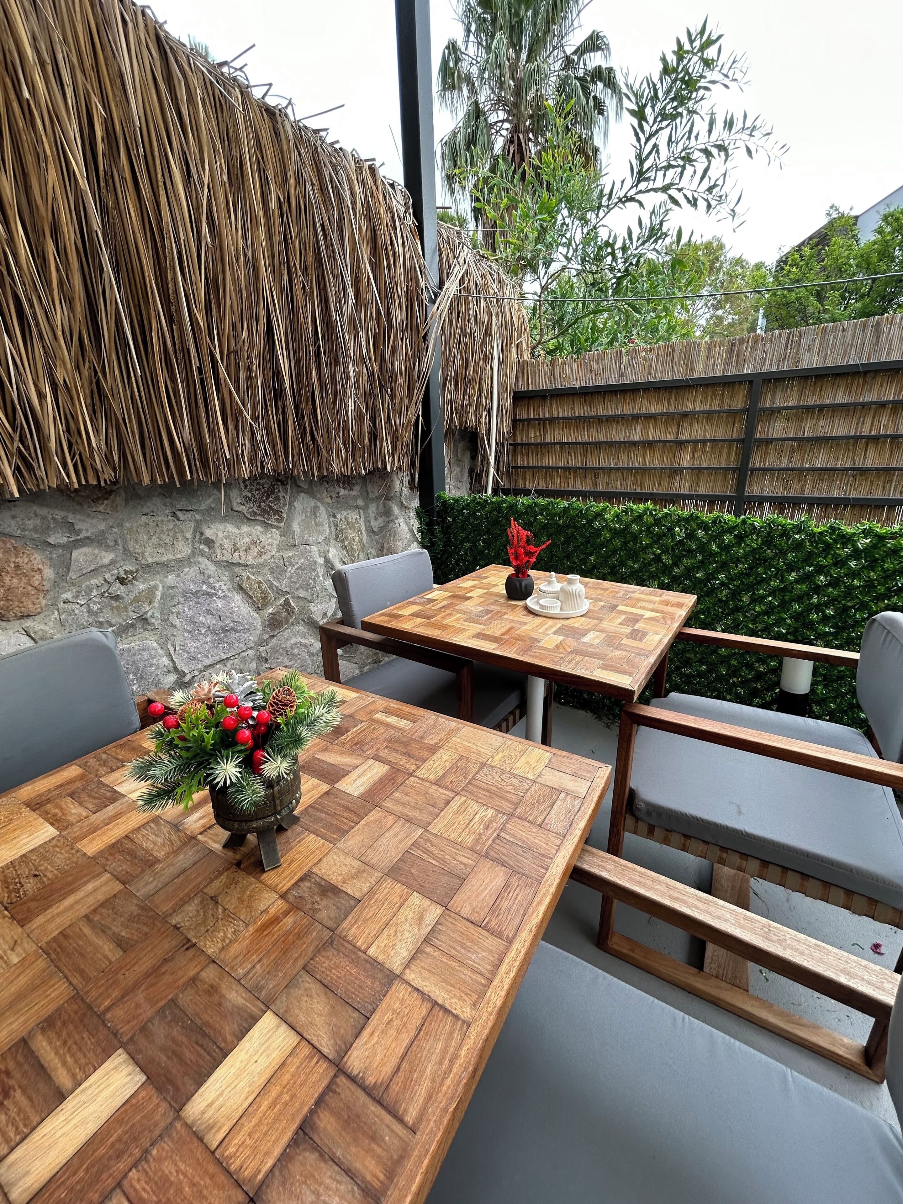 An outdoor dining area with a wooden table and chairs, a decorative centerpiece, and a thatched wall.