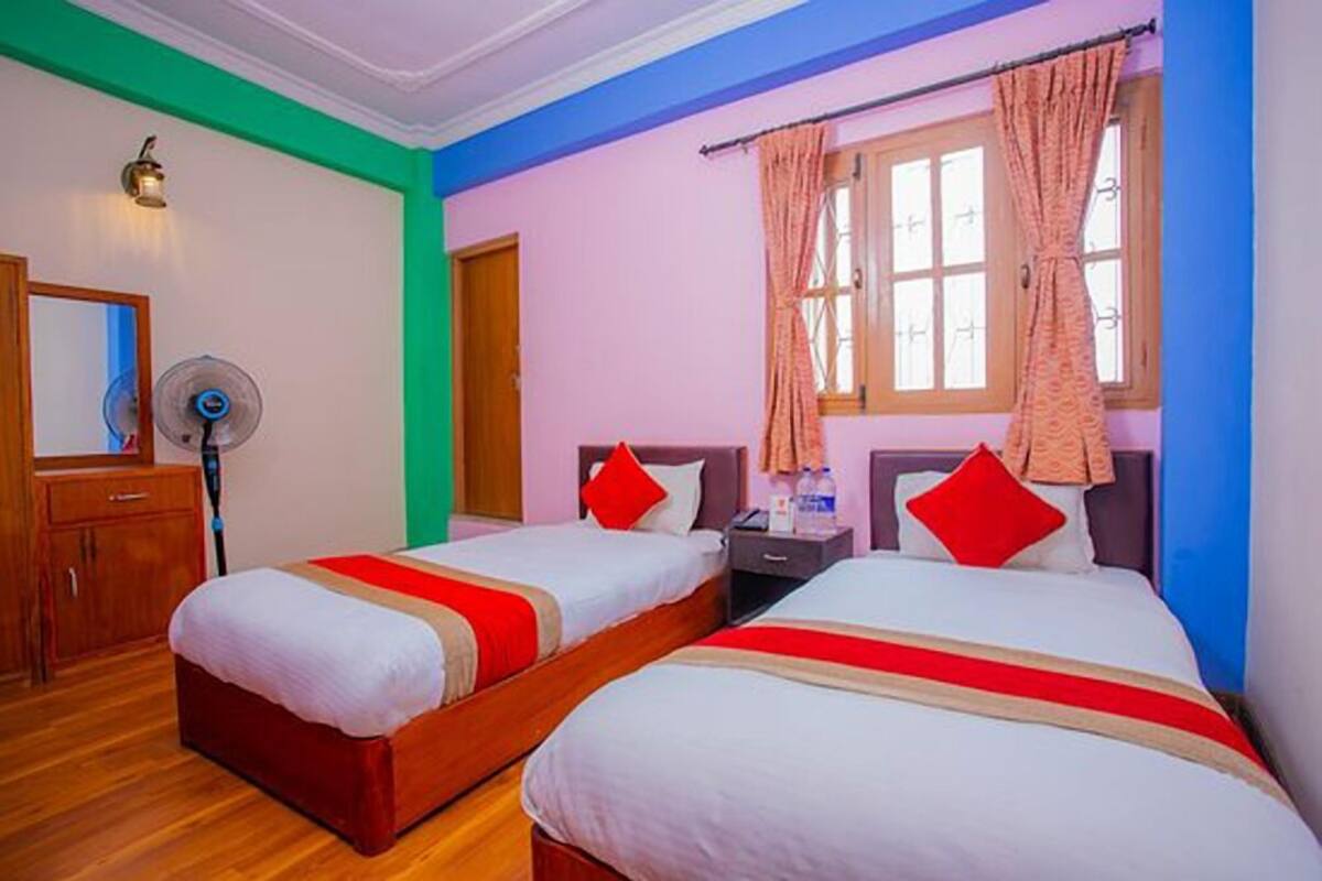 A hotel room with two single beds, a wooden wardrobe, a bedside table, a nightstand, a lamp, and a window with curtains.