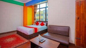 A hotel room with a bed, a sofa, a small table with a remote control, and a window with a view of buildings. - Hotel om (Kathmandu)