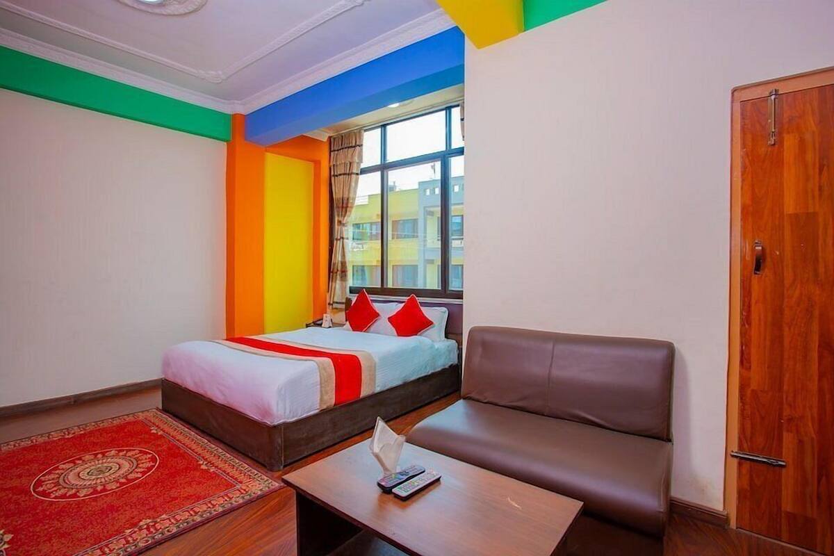 A hotel room with a bed, a sofa, a small table with a remote control, and a window with a view of buildings.