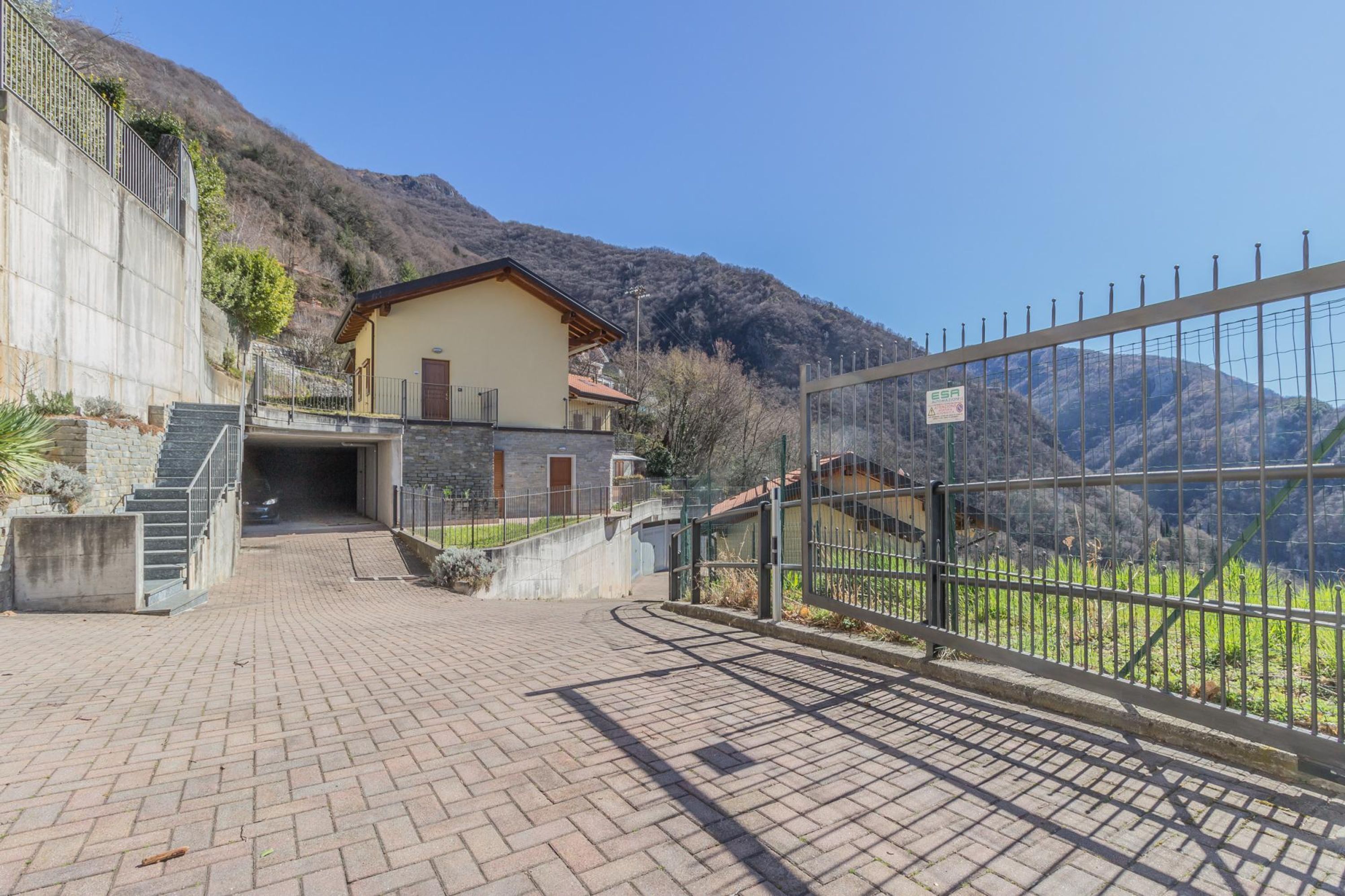 Un edificio con una scala in pietra che conduce a una porta, una zona pavimentata e un giardino recintato con montagne sullo sfondo.