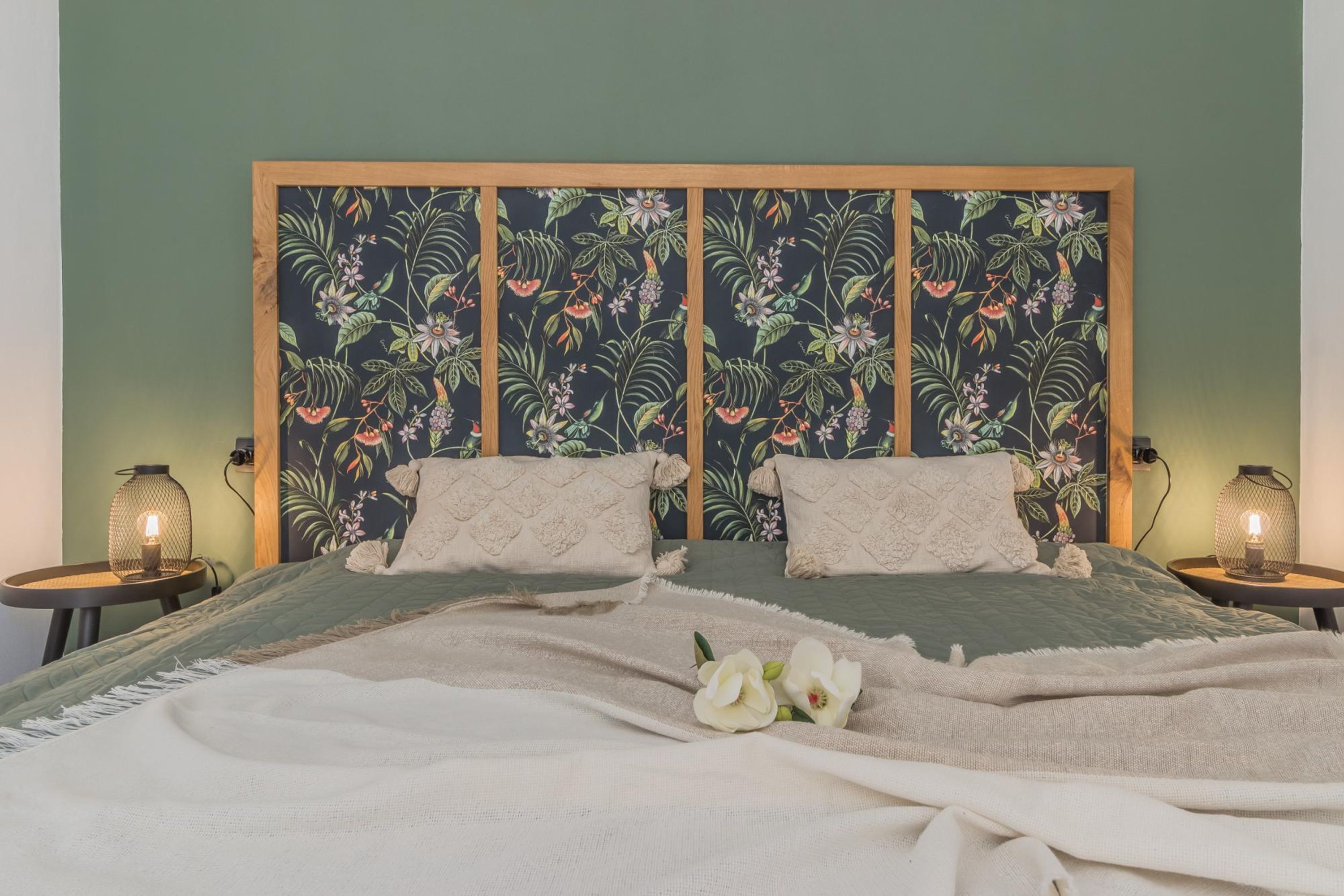 Un lit double avec une tête de lit à motifs floraux, deux tables de chevet avec des lampes et une fleur blanche décorative posée sur le lit.