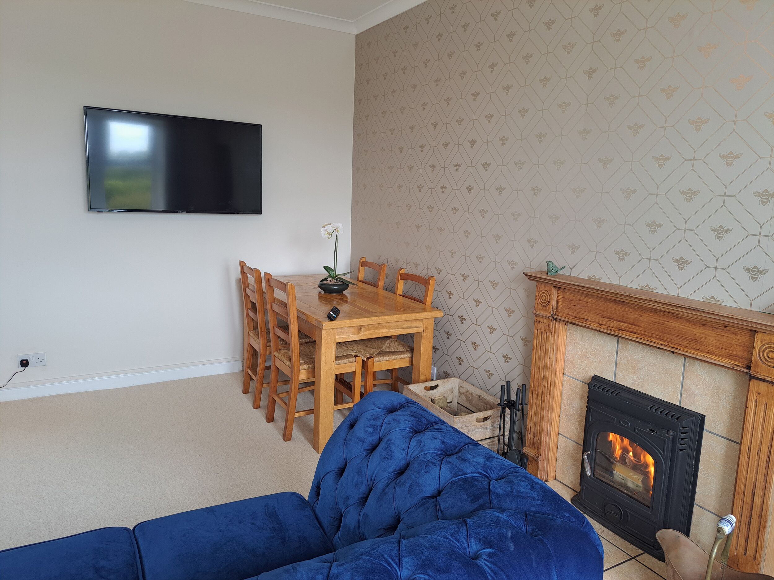 A living room with a blue sofa, a wooden dining table with chairs, a flat-screen TV mounted on the wall, and a fireplace with a tiled surround.