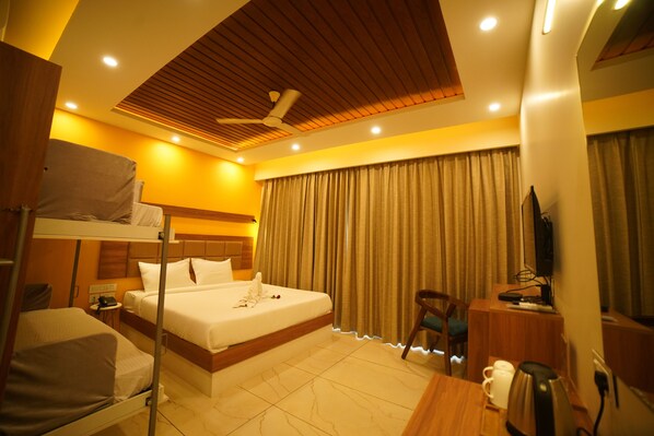 A hotel room with a bunk bed, a single bed, a desk with a chair, a television, and a coffee maker. - Aananda wellness and resorts (Beltangadi)