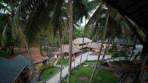 A residential area with multiple buildings, including a house with a red roof and a larger building with a grey roof. There are palm trees and a well-maintained lawn. - Aananda wellness and resorts (Beltangadi)