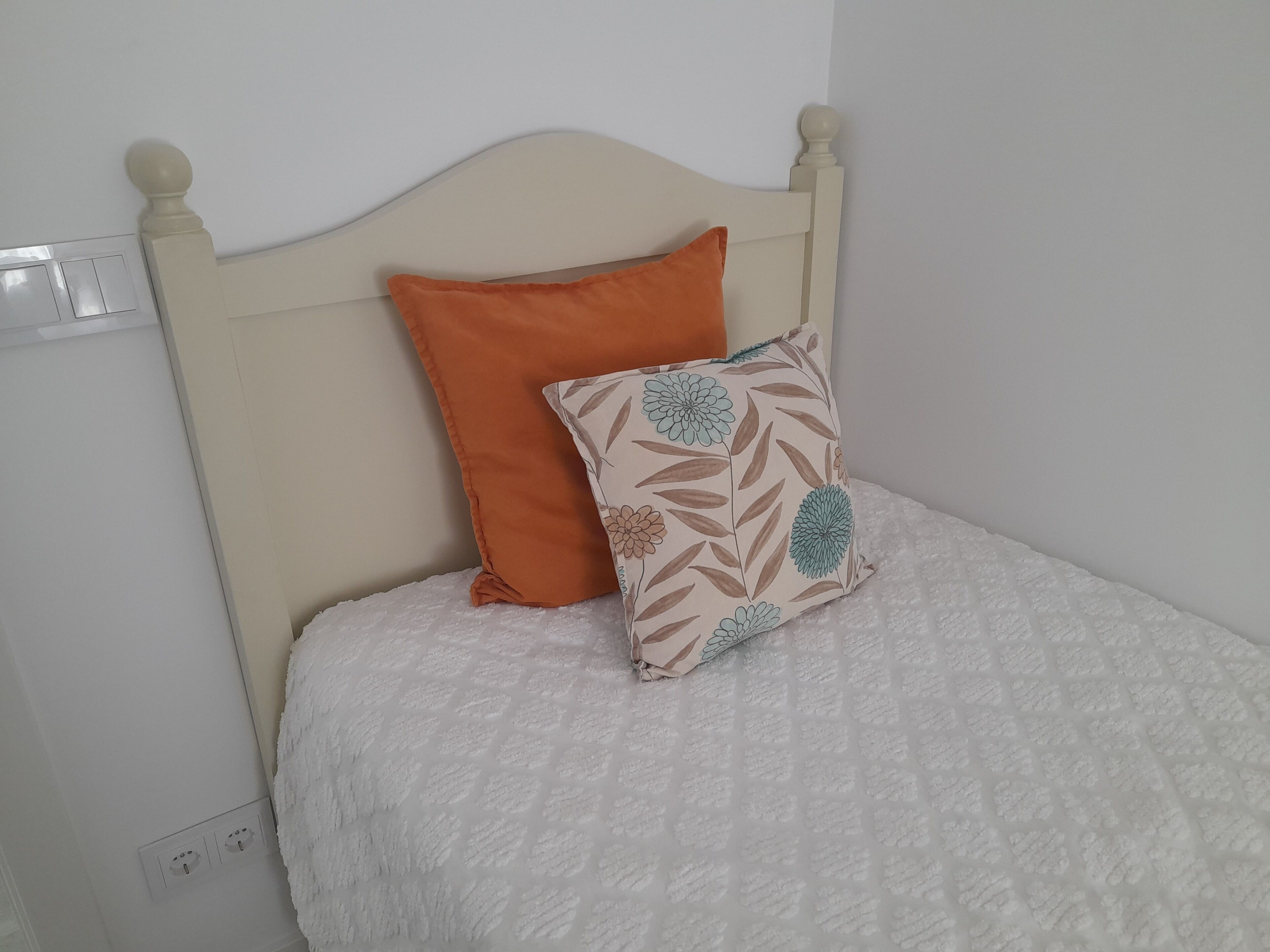 A bed with a white quilt and two pillows, one orange and one with a floral pattern.