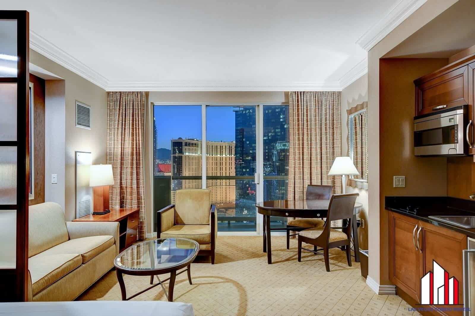 A hotel room with a city view, a sofa, a glass table, a dining table with chairs, and a kitchenette.