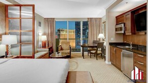 Une chambre d’hôtel dotée d’un grand lit, d’un coin salon avec une table et des chaises, d’une kitchenette et offrant une vue sur le paysage urbain la nuit.