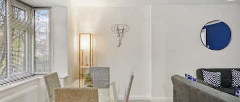 A modern living room with a glass dining table, grey upholstered chairs, a grey sofa, a wall-mounted elephant artwork, a round mirror, and a floor lamp.