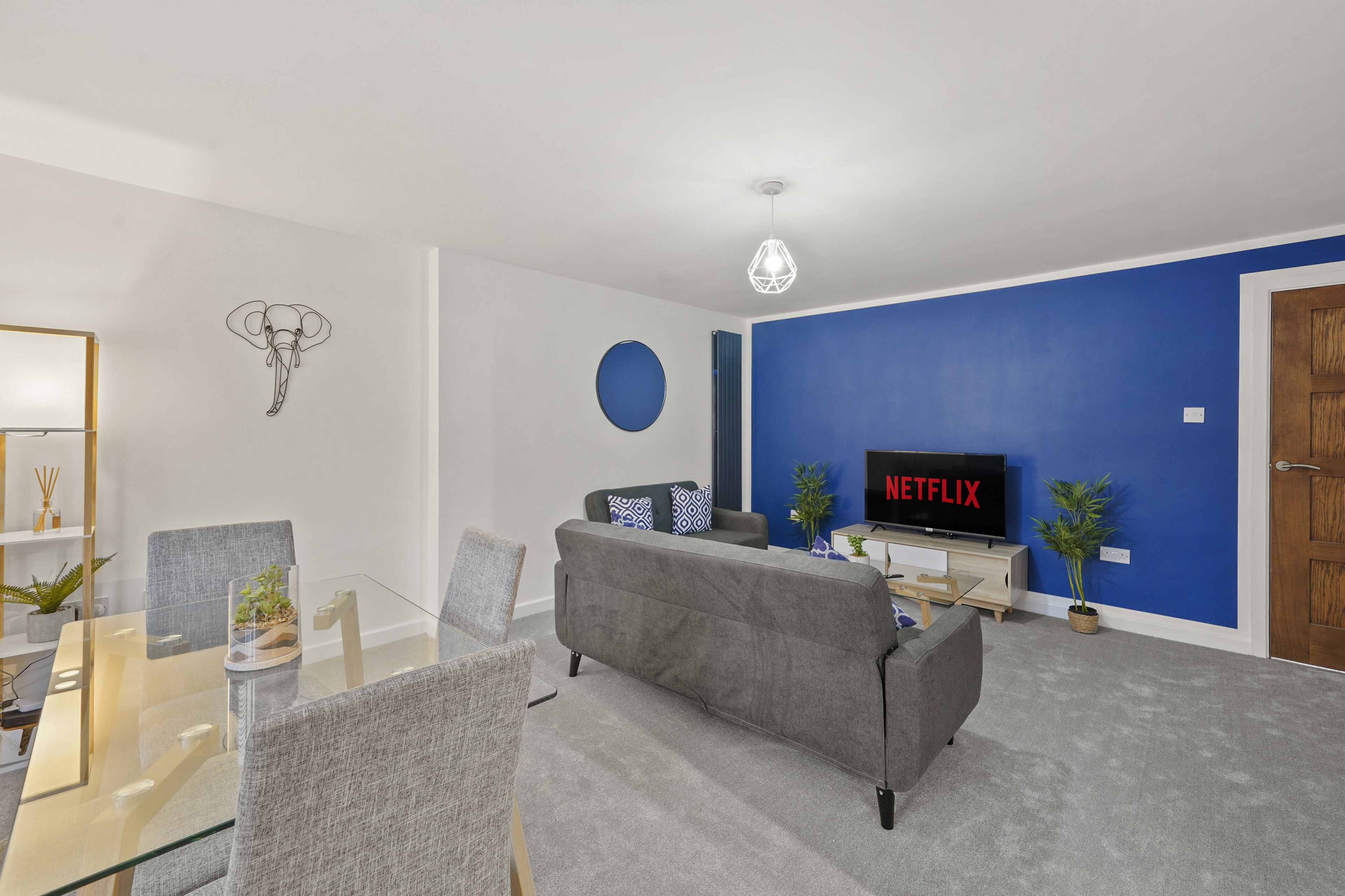 A modern living room with a glass dining table, grey sofa, and a flat-screen TV displaying Netflix.