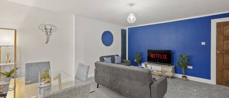 A modern living room with a glass dining table, grey sofa, and a flat-screen TV displaying Netflix.