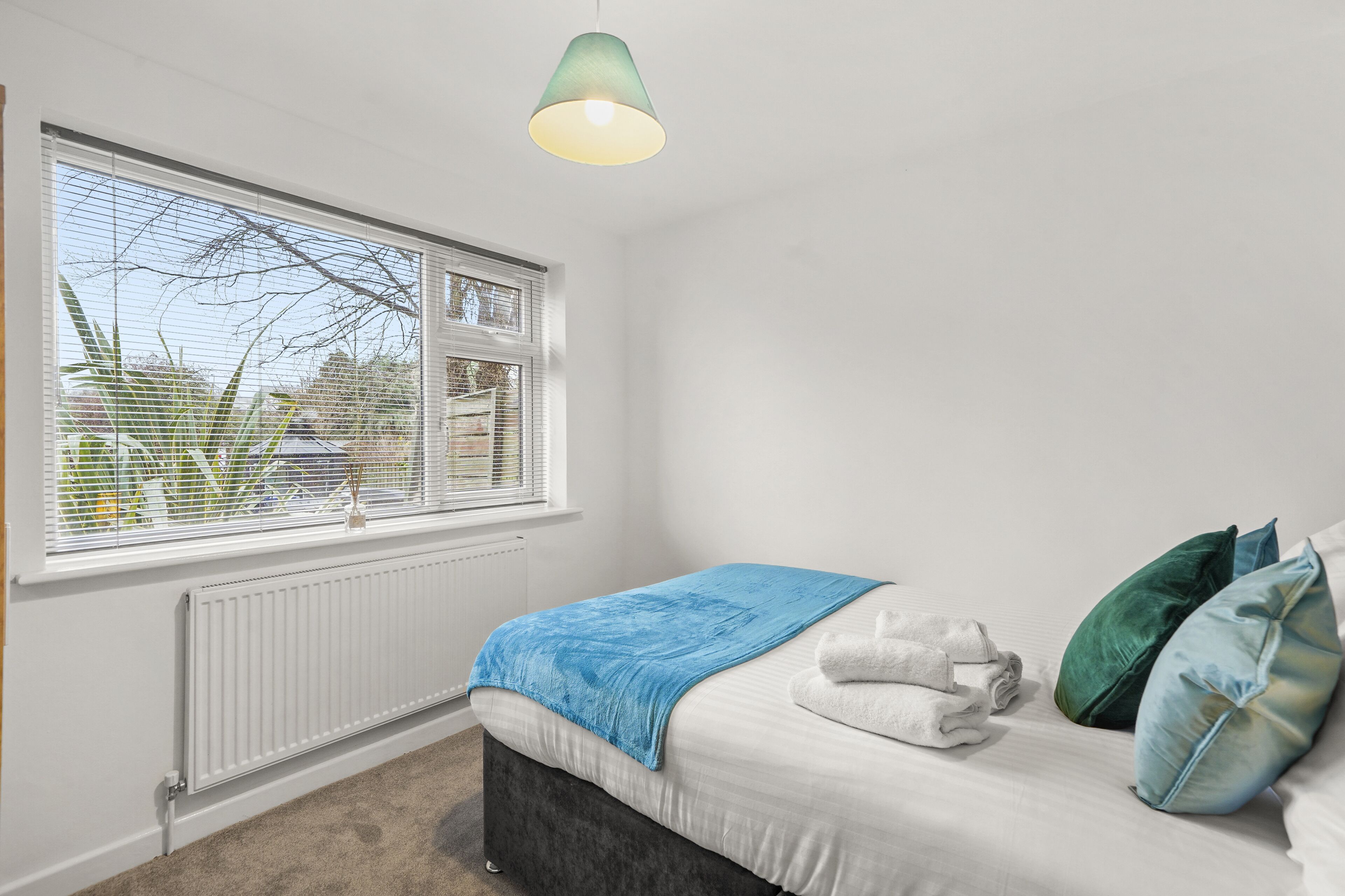 Une chambre à coucher avec un lit, une fenêtre avec des stores, un radiateur et un luminaire suspendu.