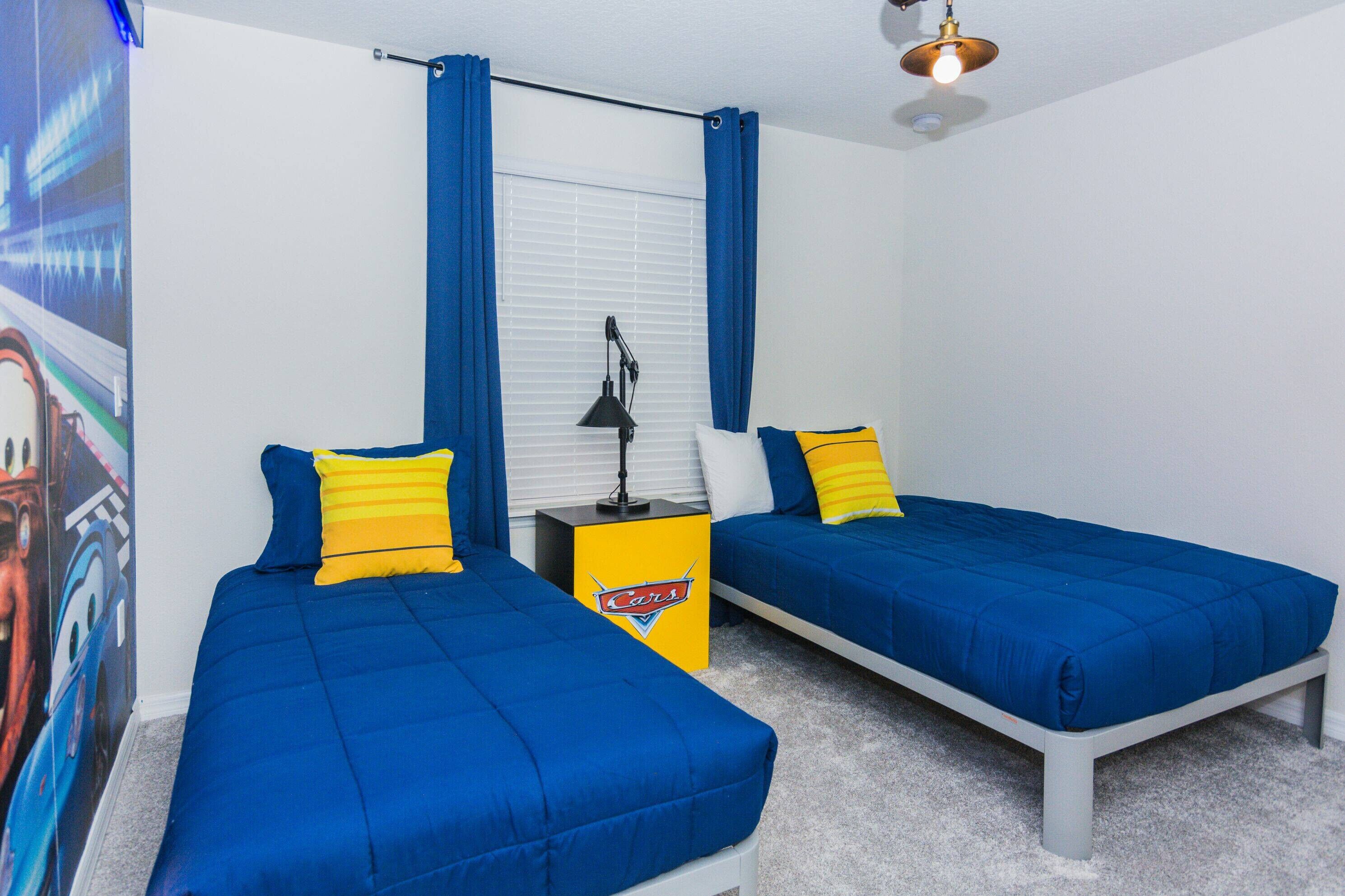 A bedroom with two single beds, each with blue bedding and yellow pillows. There is a nightstand with a lamp and a small table with a logo. The room has a window with blue curtains and a ceiling light.
