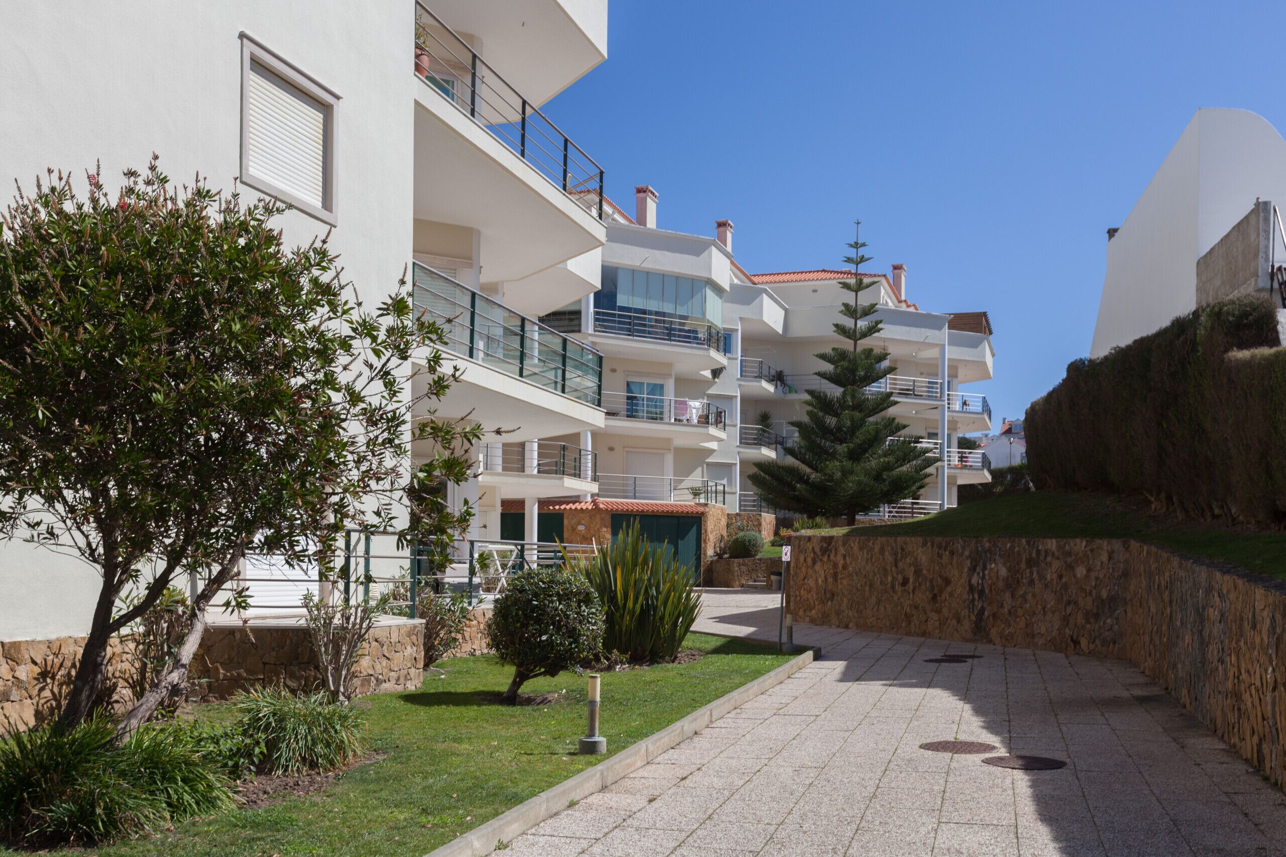 Un edificio de apartamentos moderno con varios balcones, un camino pavimentado y un jardín bien cuidado.