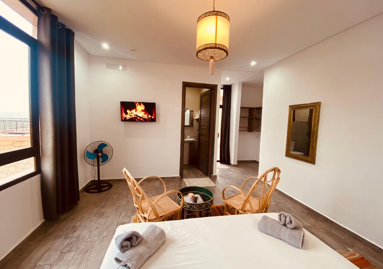 Una habitación de hotel con una cama, dos sillones de mimbre, una mesita, un ventilador y un cuadro de un fuego en la pared.