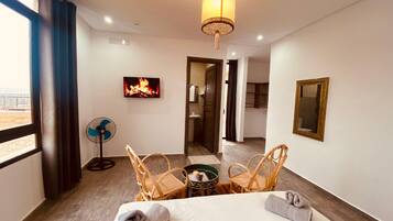 A hotel room with a bed, two wicker chairs, a small table, a fan, and a painting of a fire on the wall.