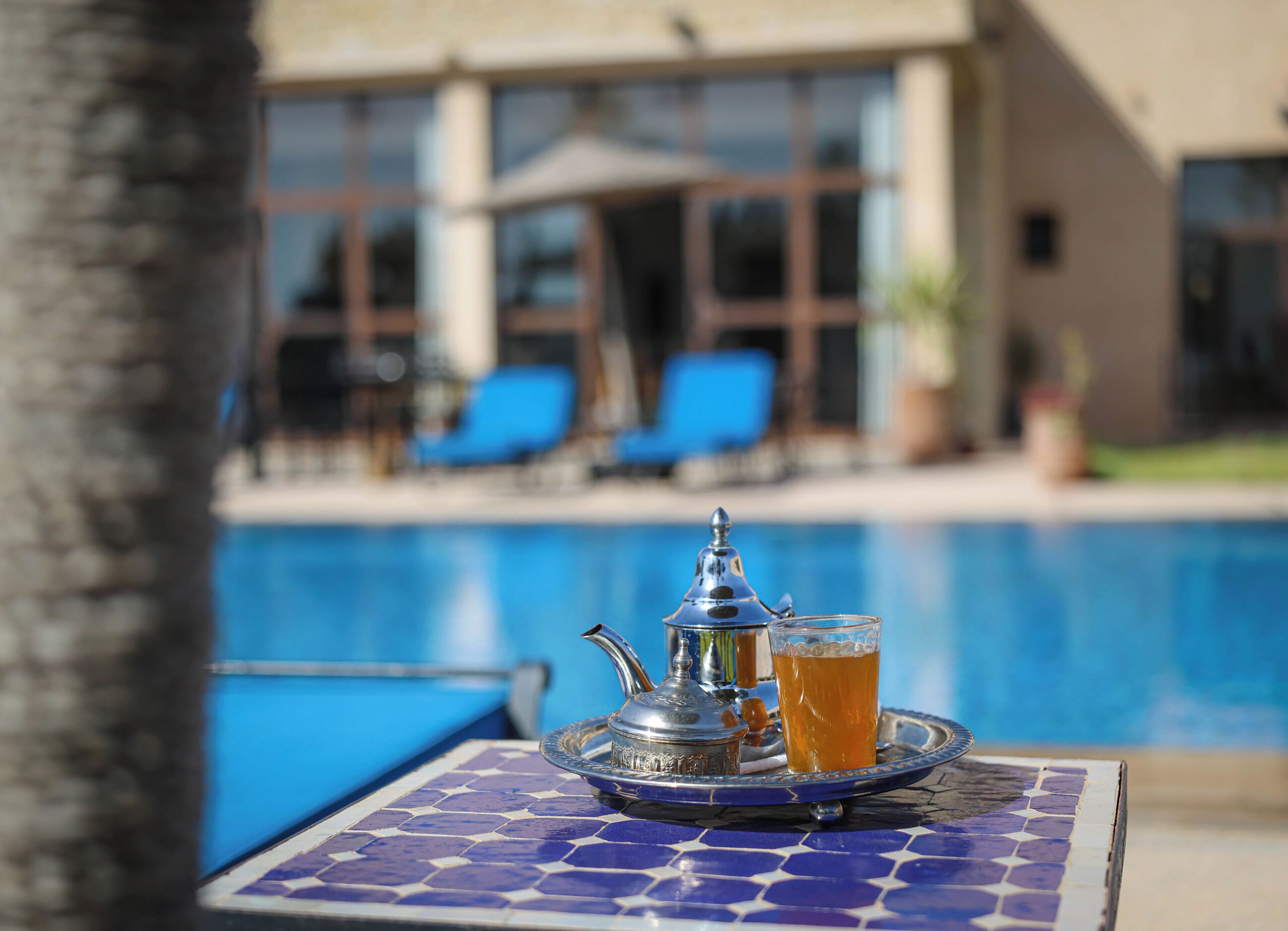 A poolside table with a teapot and a glass of tea, a pool, and a building with blue chairs in the background.