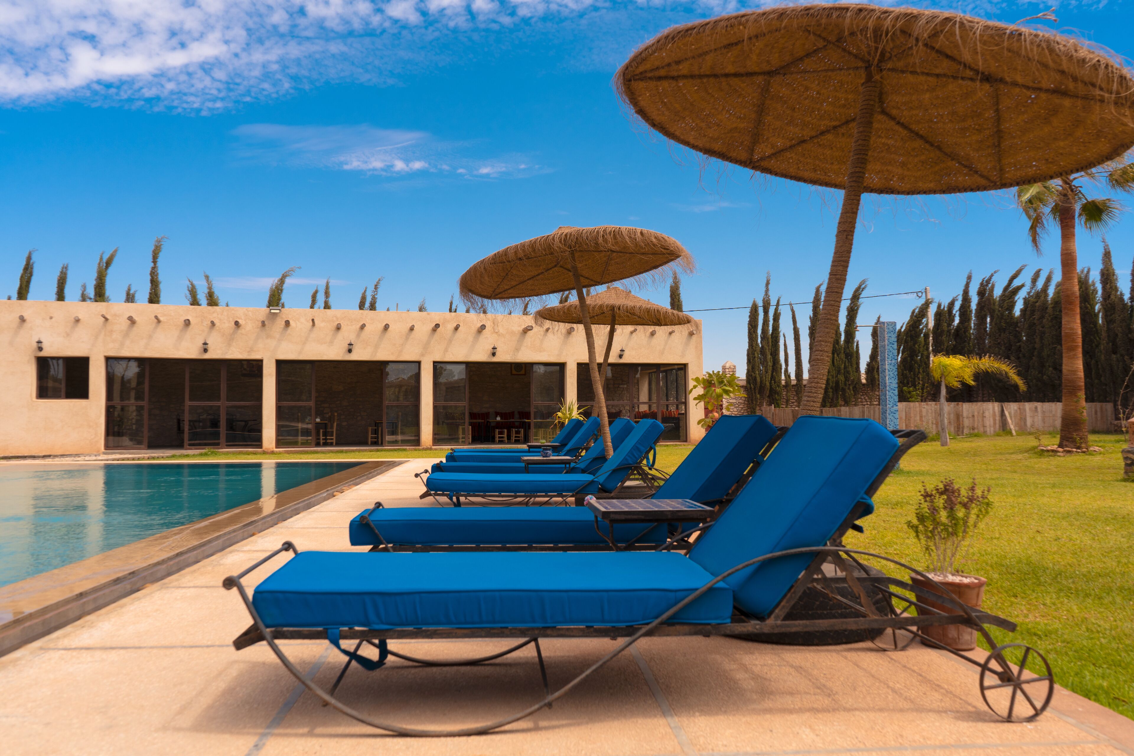 Zona junto a la piscina con sillones de descanso y sombrillas de paja, cielo azul despejado y un edificio con grandes ventanales al fondo.