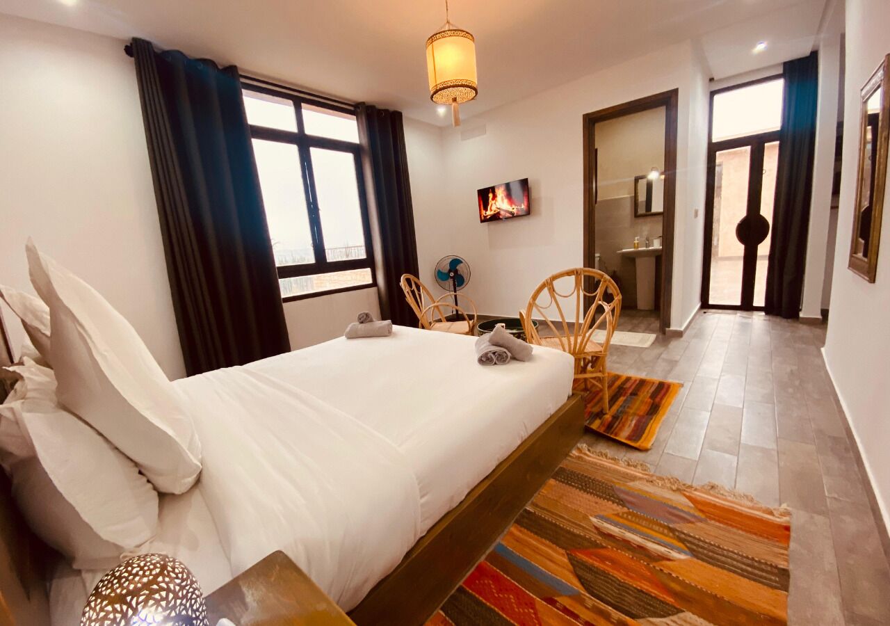 A hotel room with a bed, a fan, a chair, a rug, and a view of the outside.