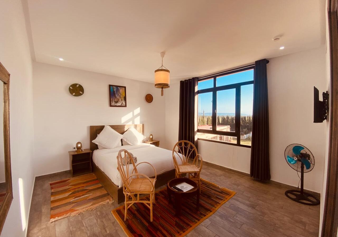 A bedroom with a large bed, two chairs, a small table, a standing fan, and a window with a view.